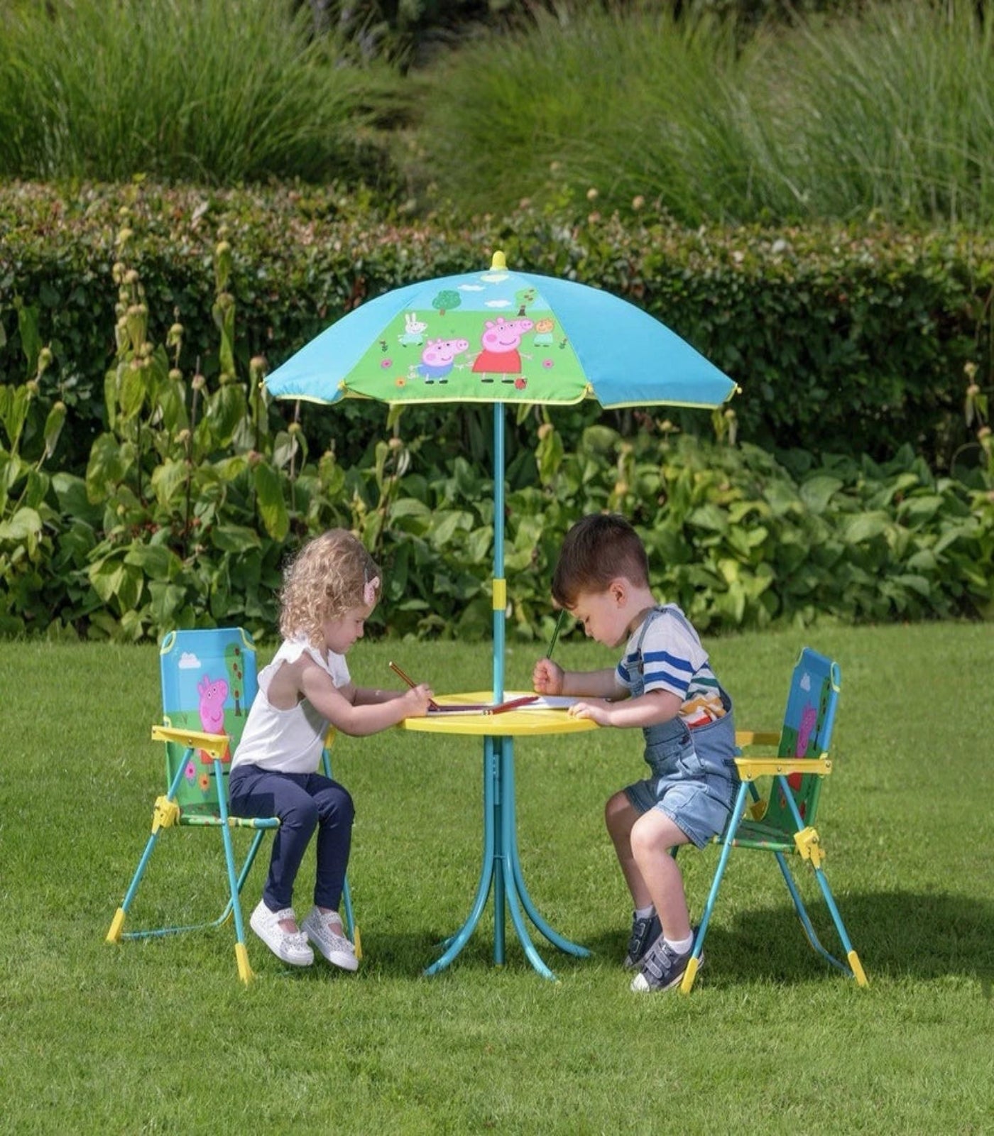 Two children sitting at a colorful outdoor table with chairs and a matching umbrella in a garden setting.