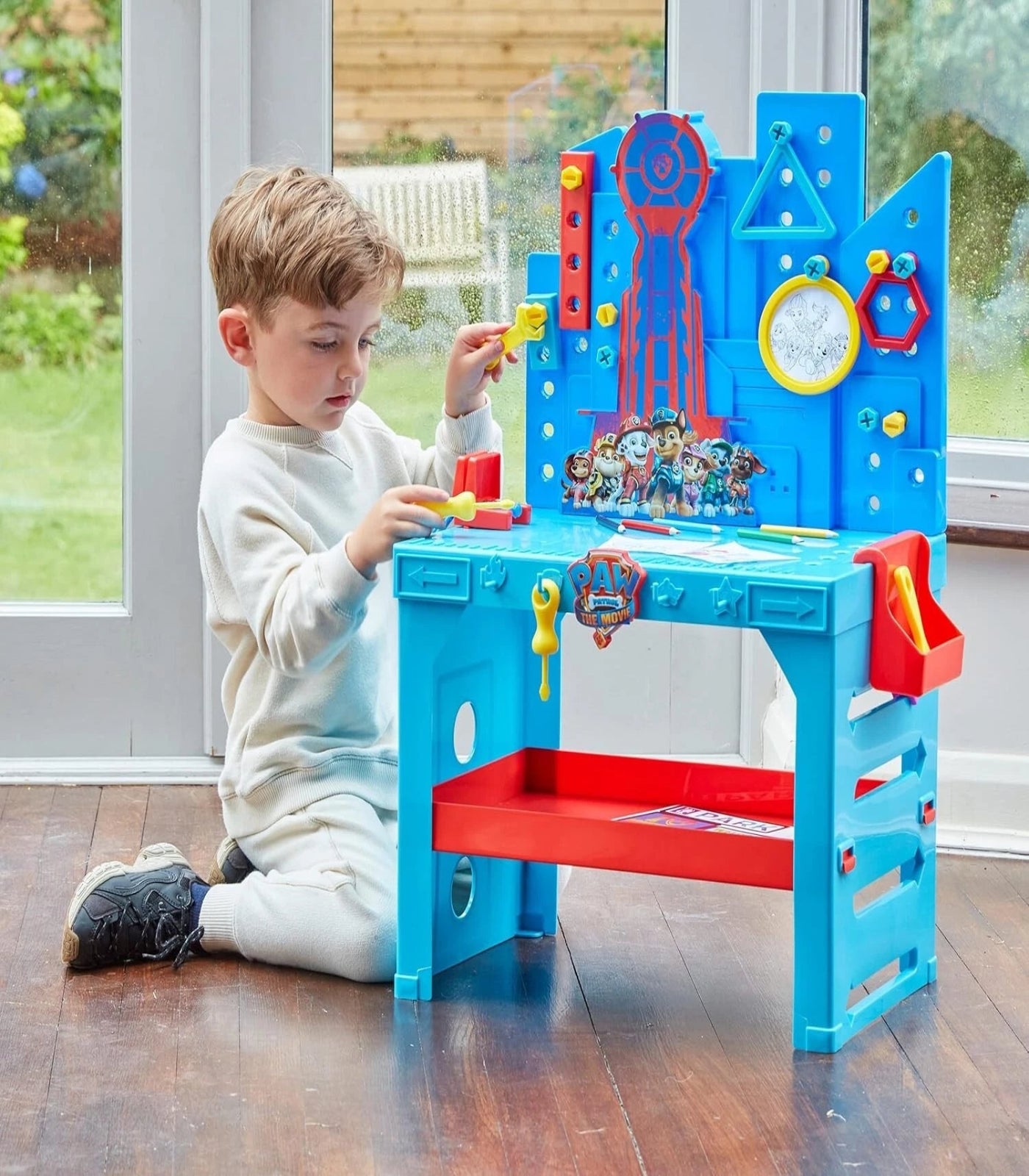 Child playing with a colorful toy desk in a room with large windows.