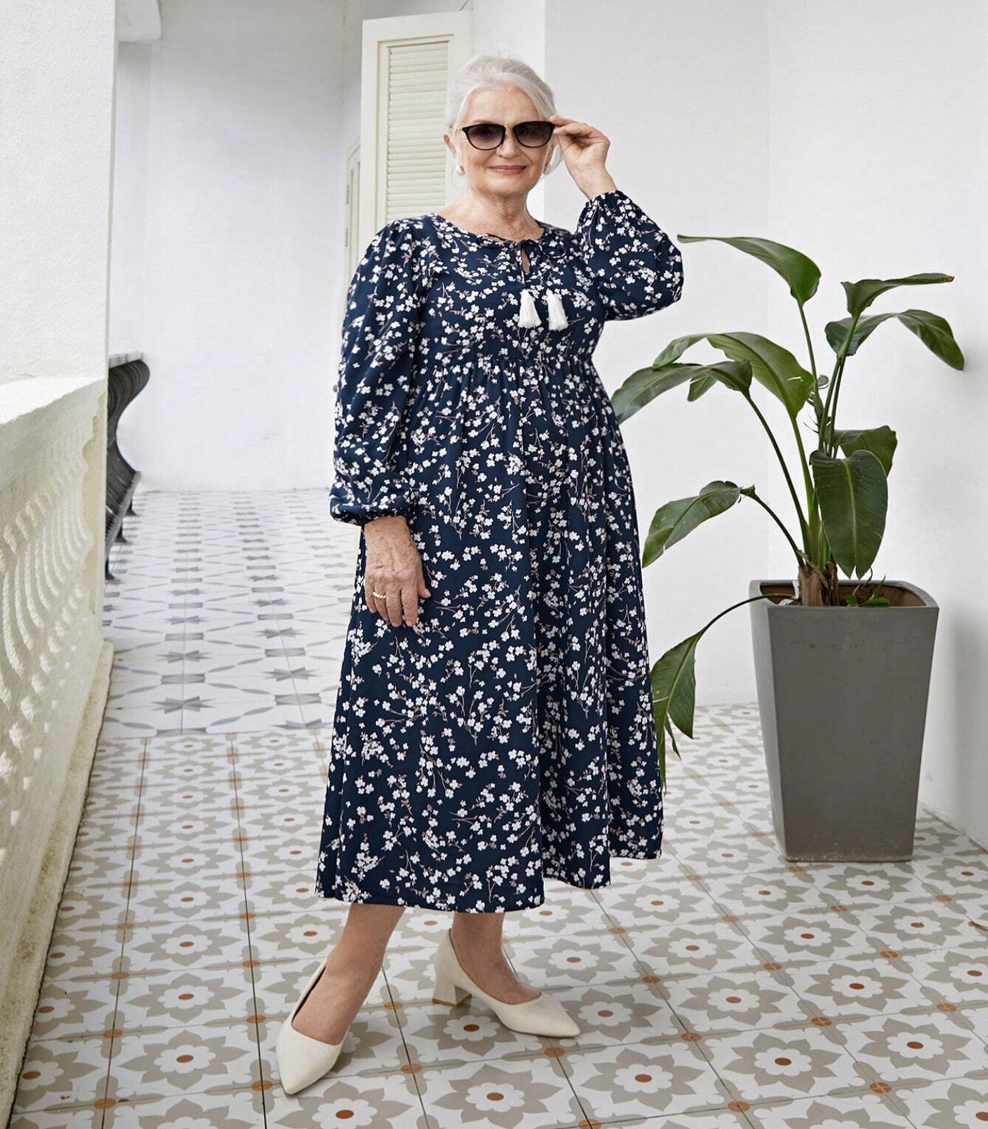 Woman in a blue floral dress standing in a room with a plant and patterned floor.