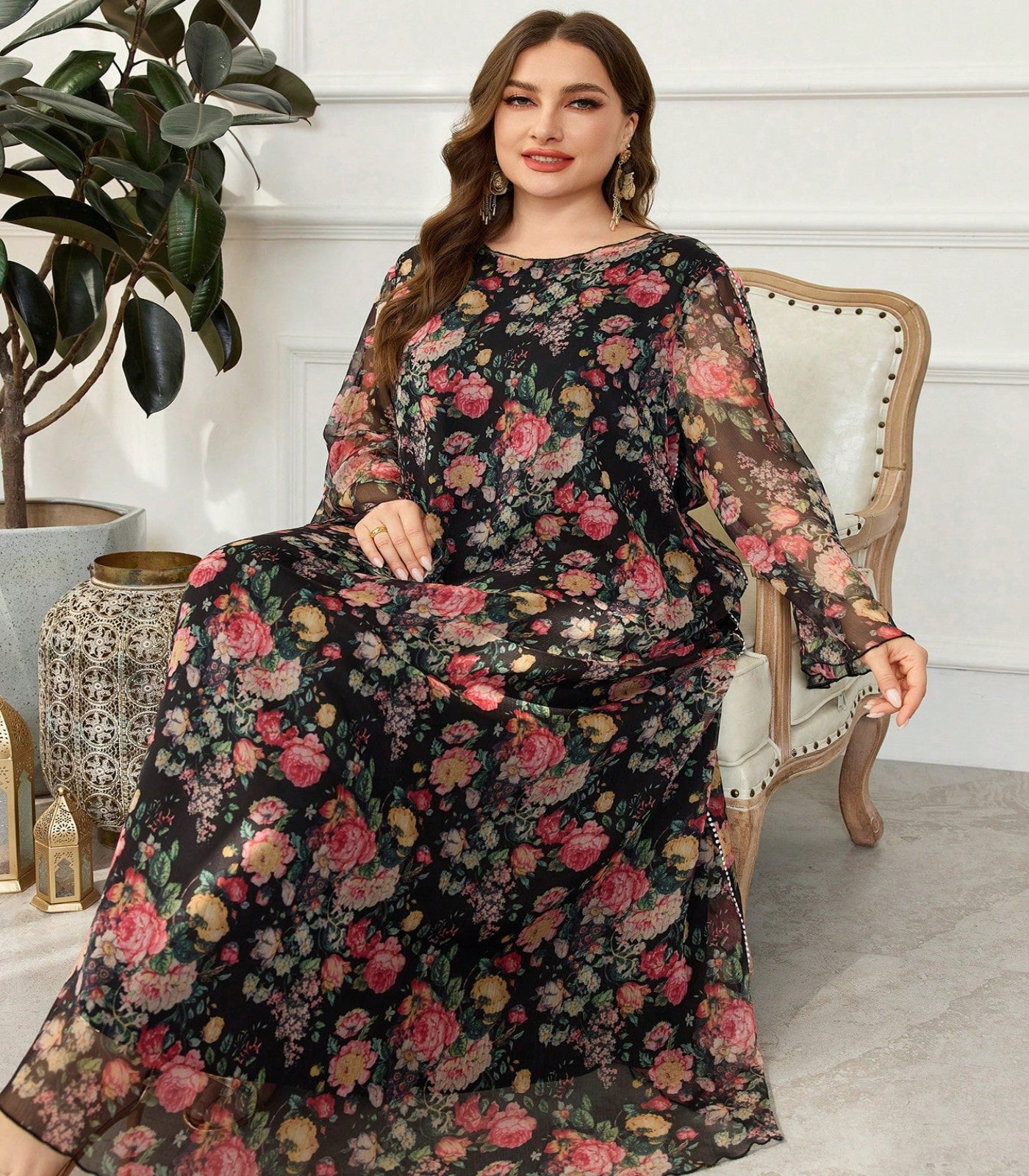Woman wearing a floral dress sitting on a chair in a room with a plant and decorative items.