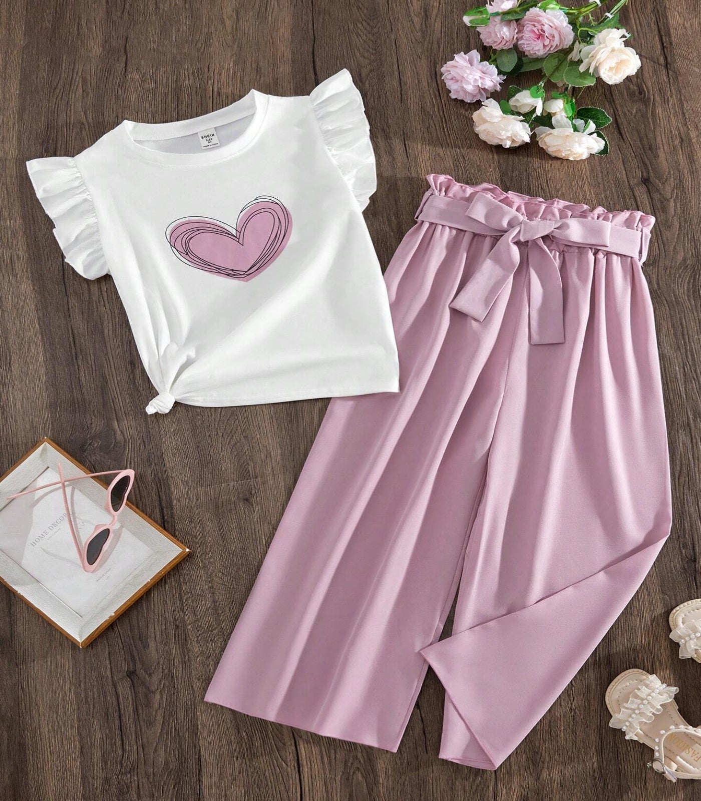 White ruffled top with pink heart design and pink pants on a wooden surface with decorative elements.