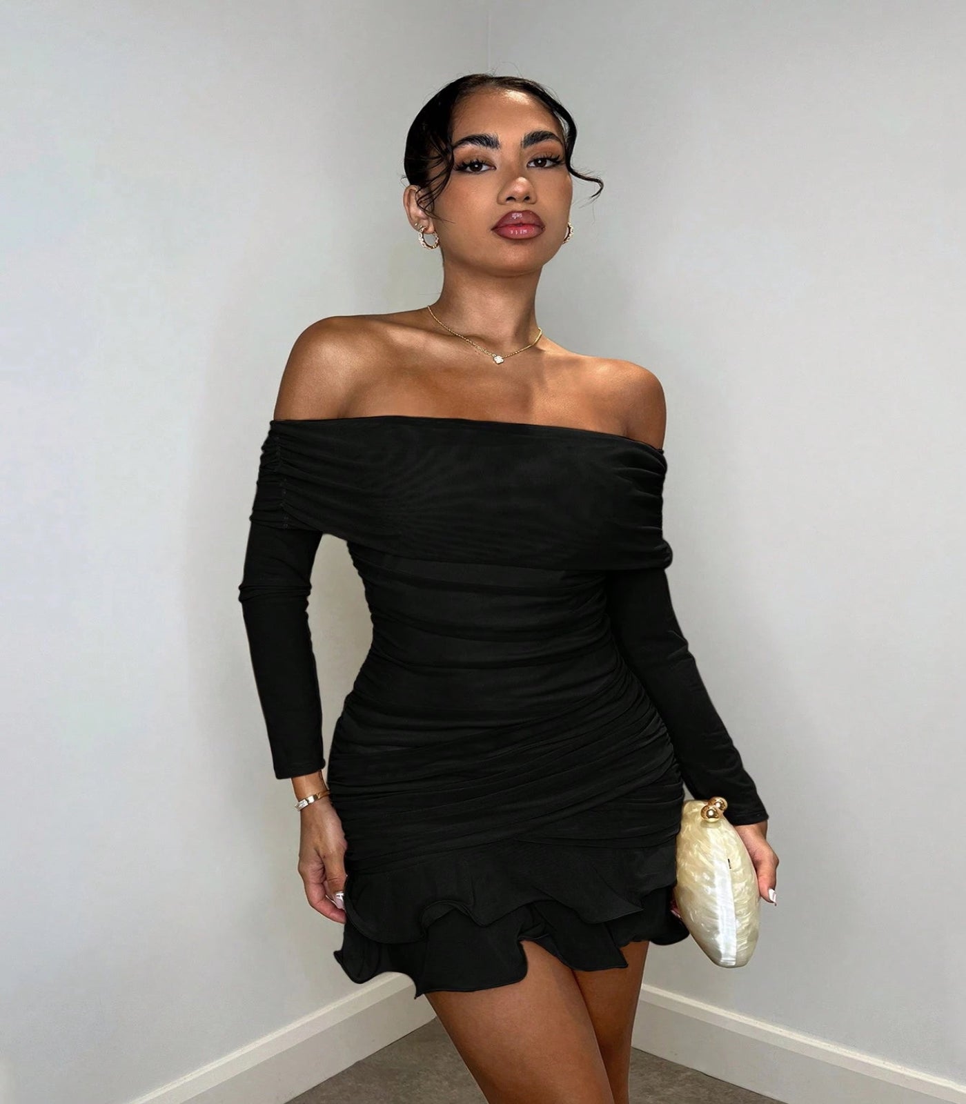 Woman wearing a black off-shoulder dress holding a gold clutch against a plain background